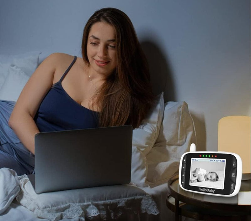 Monitor HelloBaby HB65 na stoliku przy łóżku, z czarno-białym obrazem śpiącego dziecka na ekranie.