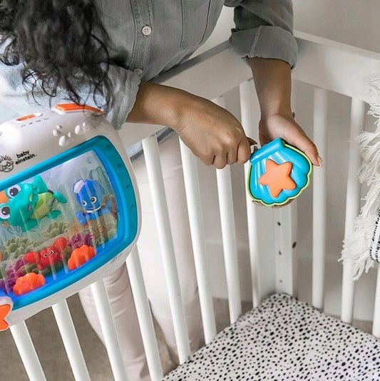 Dorosła osoba trzyma pilot z gwiazdką obok pozytywki Baby Einstein Morskie Sny przymocowanej do białego łóżeczka.