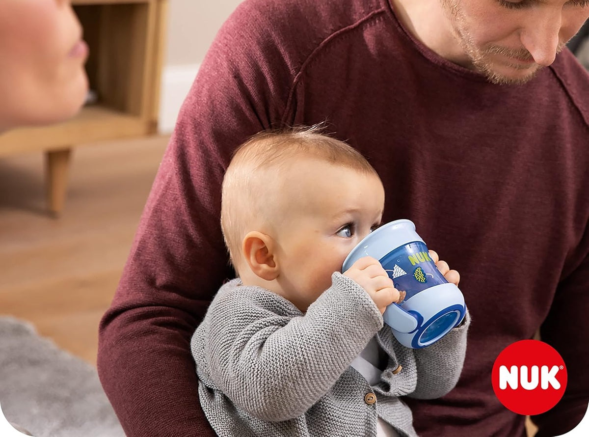 Dziecko trzymające niebieski kubek NUK Mini Magic Cup przy ustach na kolanach dorosłego.